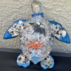May include: A decorative sea turtle figurine crafted from clear resin, featuring a blue painted head and flippers. The shell is filled with small seashells, a white coral-like structure, a small orange crab, and a black manta ray silhouette.