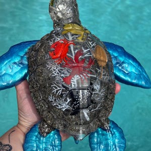 May include: A decorative sea turtle figurine featuring a clear, textured shell filled with sand and embedded with small, colorful plastic marine life figures, including a red crab, a red lobster, a gold horseshoe crab, and a black stingray.