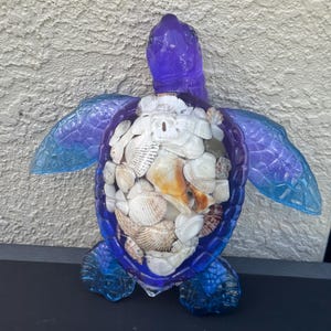 May include: A decorative sea turtle figurine crafted from translucent blue and purple resin. The turtle's shell is filled with a collection of various white, tan, and brown seashells, including a sand dollar, creating a textured mosaic effect.