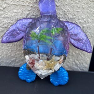 May include: A translucent purple and blue sea turtle figurine with a clear shell containing a miniature tropical scene. Inside the shell are green palm trees, white sand, various small seashells, and a tan starfish resting on the bottom.