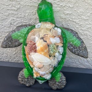 May include: A decorative sea turtle figurine featuring a translucent green glass head and flippers with a textured finish. The shell is a clear glass enclosure filled with a variety of natural seashells in shades of white, tan, and brown.