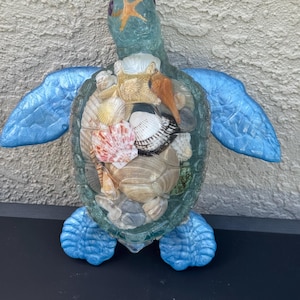 May include: A decorative sea turtle figurine with a translucent green head and blue textured flippers. The shell is a clear glass enclosure filled with a variety of natural seashells, including a tan starfish, spiral shells, and bivalves.