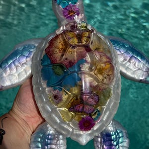 May include: A sea turtle figurine crafted from clear resin with a pearlescent white finish on the head and flippers. The shell is transparent and contains embedded dried flowers in shades of pink, purple, and blue, along with butterfly imagery.