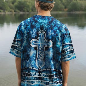 May include: A short-sleeve t-shirt featuring a blue and black tie-dye pattern with a large white cross graphic centered on the back. The word faith is written vertically in white script inside the cross. The shirt has horizontal wavy white lines.