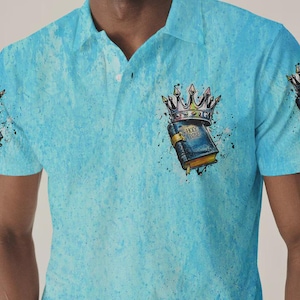 May include: A light blue polo shirt with a textured, mottled pattern. The chest features a graphic of a dark blue book labeled "HOLY BIBLE" topped with a metallic crown, surrounded by black paint splatters and ink-like artistic accents.