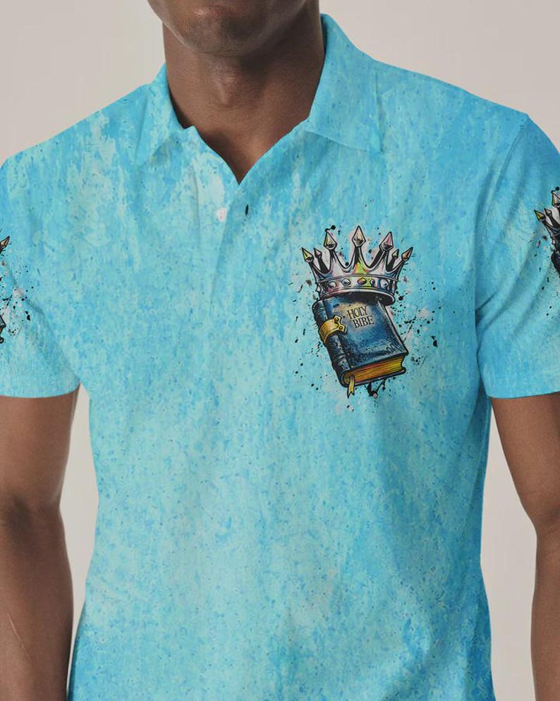 May include: A light blue polo shirt with a textured, mottled pattern. The chest features a graphic of a dark blue book labeled "HOLY BIBLE" topped with a metallic crown, surrounded by black paint splatters and ink-like artistic accents.