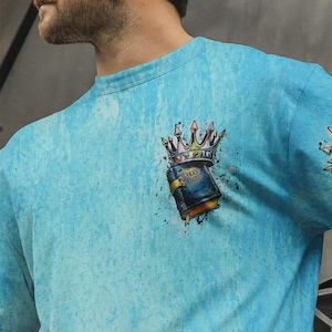May include: A light blue t-shirt with a mottled texture featuring a graphic on the chest. The graphic depicts a dark blue book labeled Holy Bible with a gold clasp, topped by a silver crown with sharp points and black ink splatter accents.