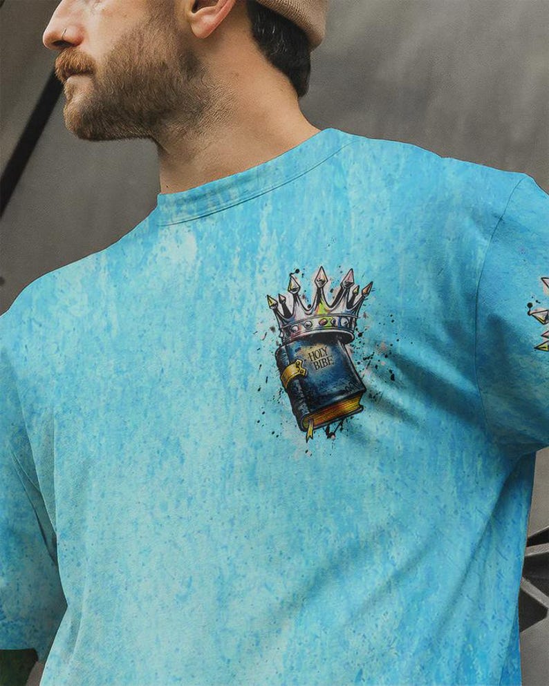 May include: A light blue t-shirt with a mottled texture featuring a graphic on the chest. The graphic depicts a dark blue book labeled Holy Bible with a gold clasp, topped by a silver crown with sharp points and black ink splatter accents.