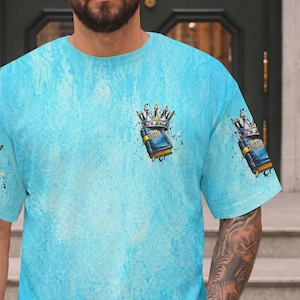 May include: A light blue t-shirt with a textured, mottled wash effect. The chest and sleeve feature a graphic illustration of a blue book labeled Holy Bible, topped with a silver crown, accented by black ink splatters and artistic shading.