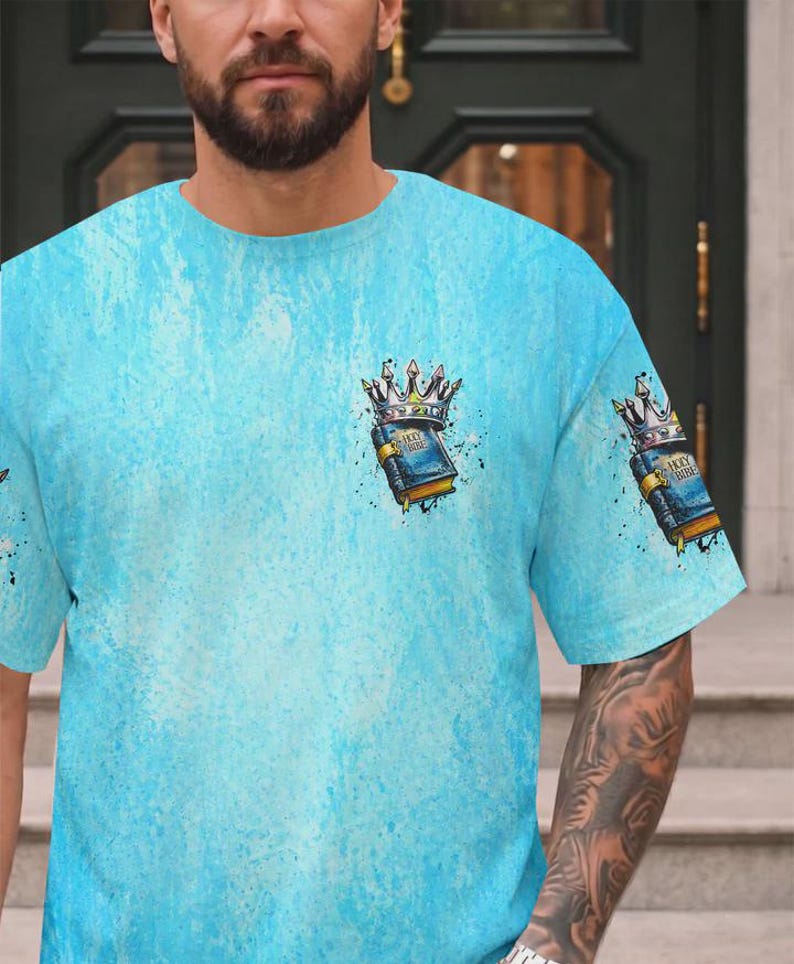 May include: A light blue t-shirt with a textured, mottled wash effect. The chest and sleeve feature a graphic illustration of a blue book labeled Holy Bible, topped with a silver crown, accented by black ink splatters and artistic shading.