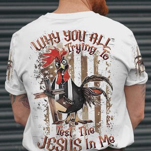 May include: A white t-shirt featuring a graphic on the back of a rooster wearing a white stole and holding a wooden cross. The design includes a distressed American flag background and the text, "WHY YOU ALL TRYING TO Test The JESUS In Me."