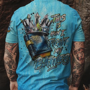 May include: A light blue t-shirt featuring a graphic print on the back. The design displays a dark blue book labeled HOLY BIBE with a gold clasp, topped by a large silver crown. The text THIS IS HOW I FIGHT MY BATTLES is printed in blue.