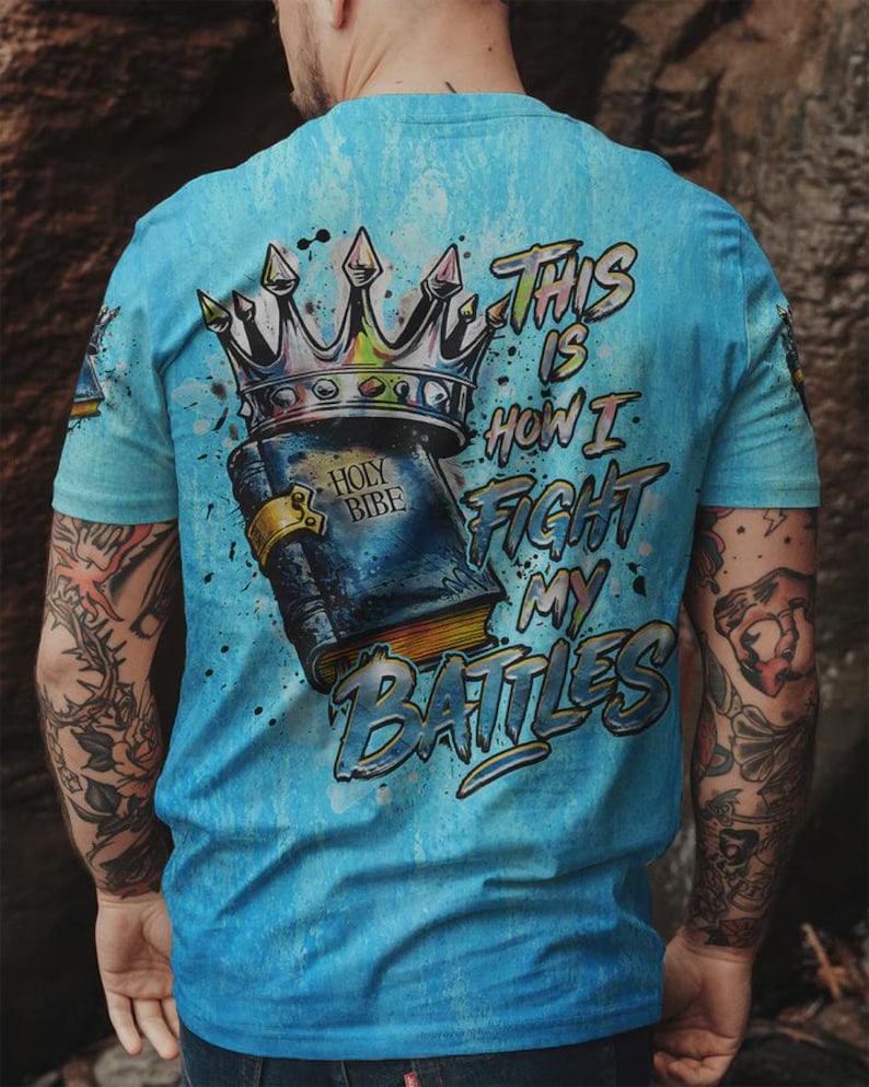 May include: A light blue t-shirt featuring a graphic print on the back. The design displays a dark blue book labeled HOLY BIBE with a gold clasp, topped by a large silver crown. The text THIS IS HOW I FIGHT MY BATTLES is printed in blue.