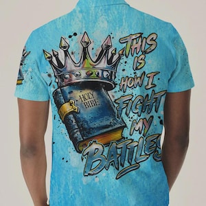 May include: A light blue short-sleeve polo shirt featuring a graphic print of a crown resting on a book labeled HOLY BIBE. The text THIS IS HOW I FIGHT MY BATTLES is printed in a stylized font next to the book, with paint splatter accents.
