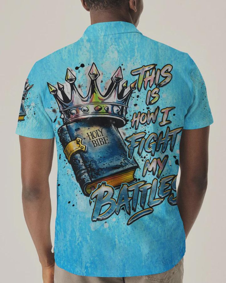 May include: A light blue short-sleeve polo shirt featuring a graphic print of a crown resting on a book labeled HOLY BIBE. The text THIS IS HOW I FIGHT MY BATTLES is printed in a stylized font next to the book, with paint splatter accents.