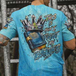 May include: A light blue t-shirt featuring a graphic print of a book labeled Holy Bible topped with a crown. The text reads This Is How I Fight My Battles. The design includes paint splatter effects and smaller crown graphics on the sleeves.
