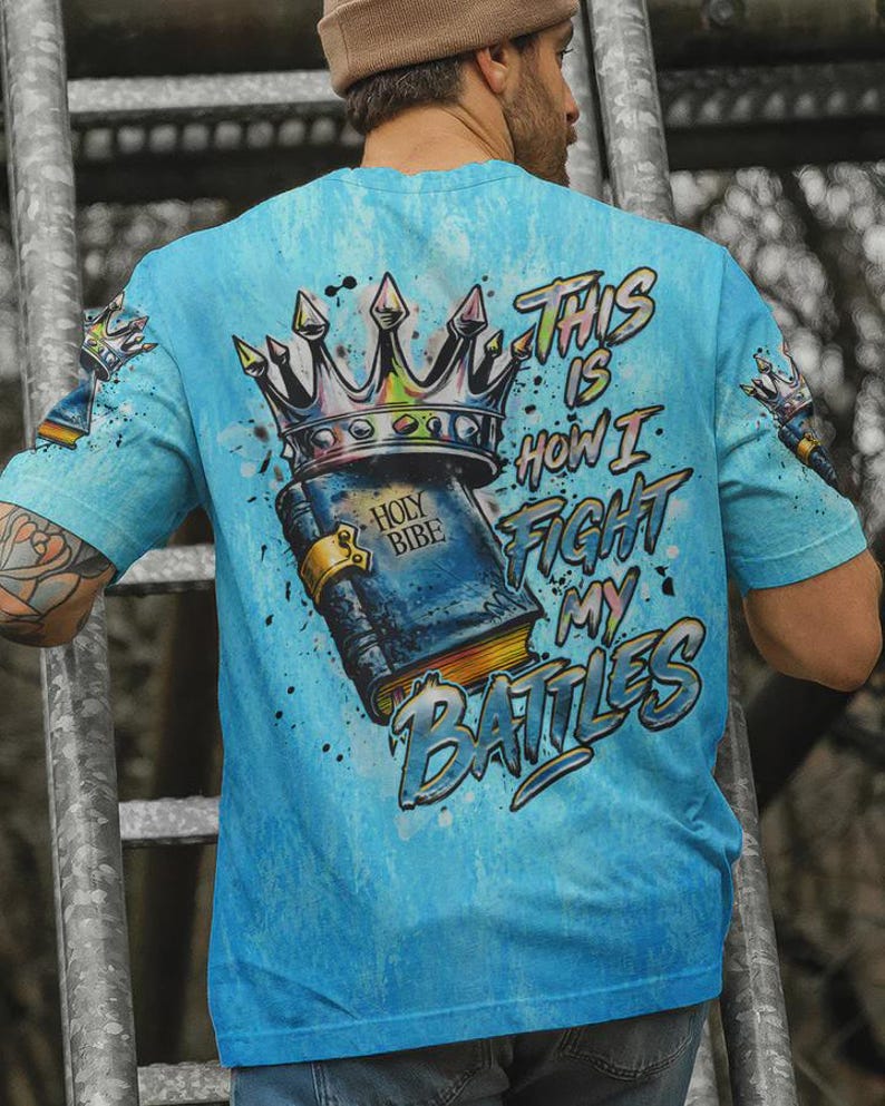 May include: A light blue t-shirt featuring a graphic print of a book labeled Holy Bible topped with a crown. The text reads This Is How I Fight My Battles. The design includes paint splatter effects and smaller crown graphics on the sleeves.