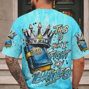 May include: A light blue t-shirt featuring a graphic print of a crown resting on a book labeled Holy Bibe. The text This Is How I Fight My Battles is printed in a stylized font. Smaller versions of the crown and book appear on the sleeves.