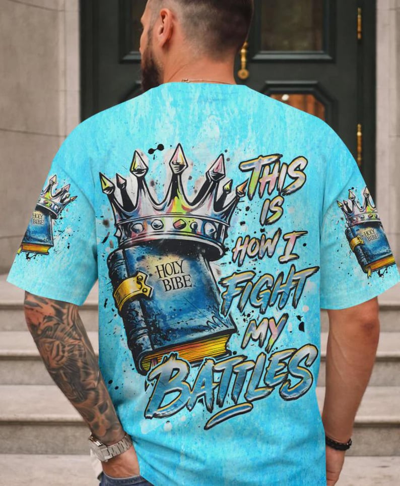 May include: A light blue t-shirt featuring a graphic print of a crown resting on a book labeled Holy Bibe. The text This Is How I Fight My Battles is printed in a stylized font. Smaller versions of the crown and book appear on the sleeves.