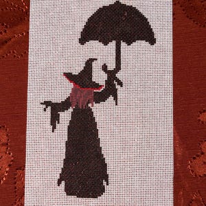 May include: A cross-stitch pattern on white fabric featuring a silhouette of a figure wearing a pointed hat and a long dress, holding a large umbrella. The figure has dark hair with a red accent, and the entire design is rendered in black thread.