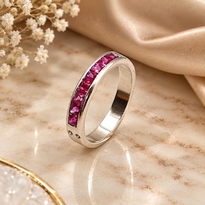 Può includere: Un anello a fascia color argento con incastonatura a binario che ospita una fila di gemme rosa acceso dal taglio quadrato. La fascia in metallo presenta una finitura liscia e lucida, con due piccoli dettagli circolari alla base.