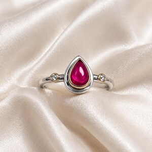 Könnte beinhalten: Ein Silberring mit einem zentralen, birnenförmigen, dunkelrosa Cabochon-Edelstein in einer goldfarbenen Fassung. Die Ringschiene ist auf beiden Seiten mit zwei kleinen, runden, klaren Steinen besetzt, die in silbernen Fassungen mit Goldakzenten sitzen.