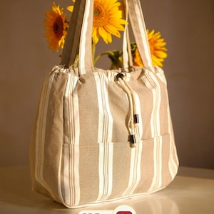 Peut inclure: Un sac fourre-tout à rayures verticales beiges et blanches avec deux bandoulières, une fermeture à cordon avec des cordons crème et des embouts noirs, et une poche extérieure. Le sac est présenté avec des tournesols jaunes.