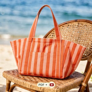 Puede incluir: Un bolso tote con rayas verticales en tonos alternos de naranja y melocotón claro. El bolso tiene dos asas largas de tela, una abertura superior ancha y un bolsillo frontal rectangular centrado entre las dos correas verticales.