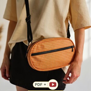 Puede incluir: Un bolso bandolera ovalado de color naranja con un patrón de cuadrícula blanca, con cierre de cremallera negra y correa negra ajustable. La imagen incluye el texto PDF + y +BONUS DRAWSTRING POUCH con un icono de botón de reproducción rojo.
