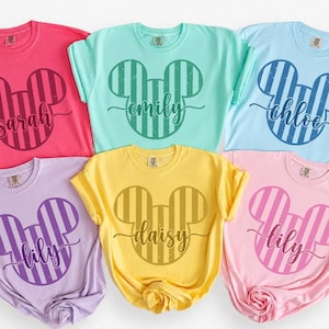 K&ouml;nnte beinhalten: Sechs kurz&auml;rmelige T-Shirts mit Rundhalsausschnitt in verschiedenen Pastellfarben wie Pink, Mintgr&uuml;n, Hellblau, Lavendel, Gelb und Hellrosa. Jedes T-Shirt zeigt eine gro&szlig;e, gestreifte Mickey-Maus-Kopf-Silhouette mit einem Namen in Schreibschrift.