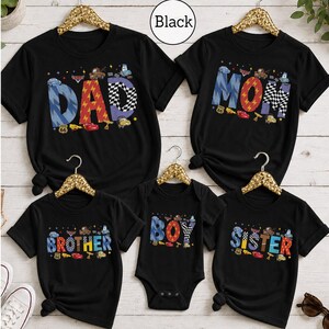 May include: Five black t-shirts and one black bodysuit featuring colorful graphic lettering. The shirts display the words DAD, MOM, BROTHER, SISTER, and BOY. Each letter is decorated with cartoon car characters, checkered patterns, and lightning bolts.