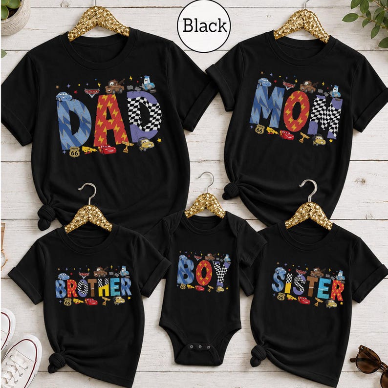 May include: Five black t-shirts and one black bodysuit featuring colorful graphic lettering. The shirts display the words DAD, MOM, BROTHER, SISTER, and BOY. Each letter is decorated with cartoon car characters, checkered patterns, and lightning bolts.