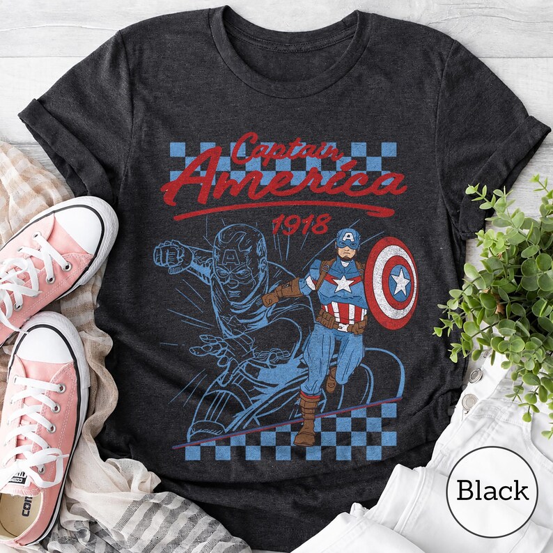 Pu&ograve; includere: Una t-shirt a maniche corte grigio antracite con una stampa grafica di Captain America. Il design include il testo Captain America 1918, un motivo a scacchi blu e bianchi e l'illustrazione di uno scudo rosso, bianco e blu.