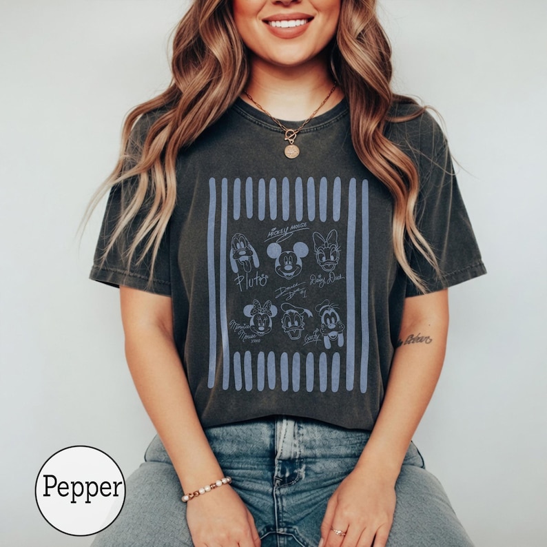 May include: A charcoal gray short-sleeve t-shirt featuring a light blue rectangular graphic with vertical stripes. The graphic contains line art illustrations of six cartoon characters with their names written below each, including Pluto, Mickey, Daisy, Minnie, Donald, and Goofy.