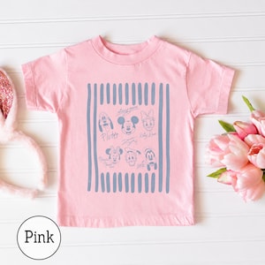 May include: A light pink short-sleeve t-shirt featuring a central rectangular graphic with vertical blue stripes. The graphic contains line-art illustrations of Mickey Mouse, Minnie Mouse, Pluto, Donald Duck, Daisy Duck, and Goofy with their names.