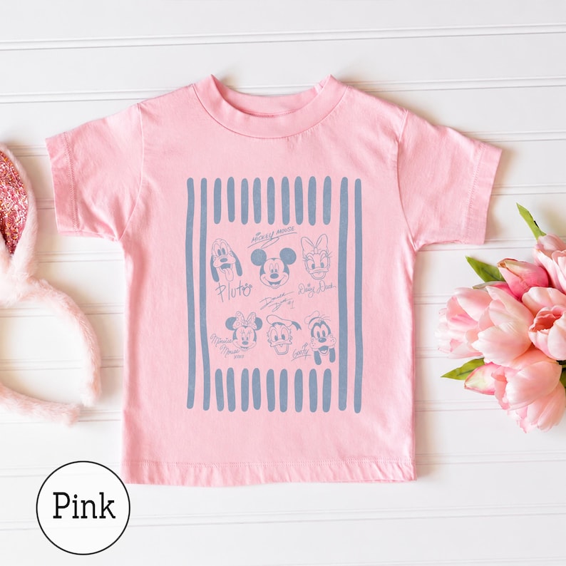 May include: A light pink short-sleeve t-shirt featuring a central rectangular graphic with vertical blue stripes. The graphic contains line-art illustrations of Mickey Mouse, Minnie Mouse, Pluto, Donald Duck, Daisy Duck, and Goofy with their names.