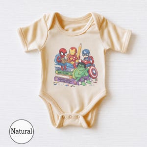 Puede incluir: Un body de manga corta en color beige claro con un estampado gr&aacute;fico de superh&eacute;roes de dibujos animados, incluyendo a Spider-Man, Iron Man, Captain America y Hulk, sentados sobre una pila de libros titulados Super Physics, World History y Avengers Assemble.