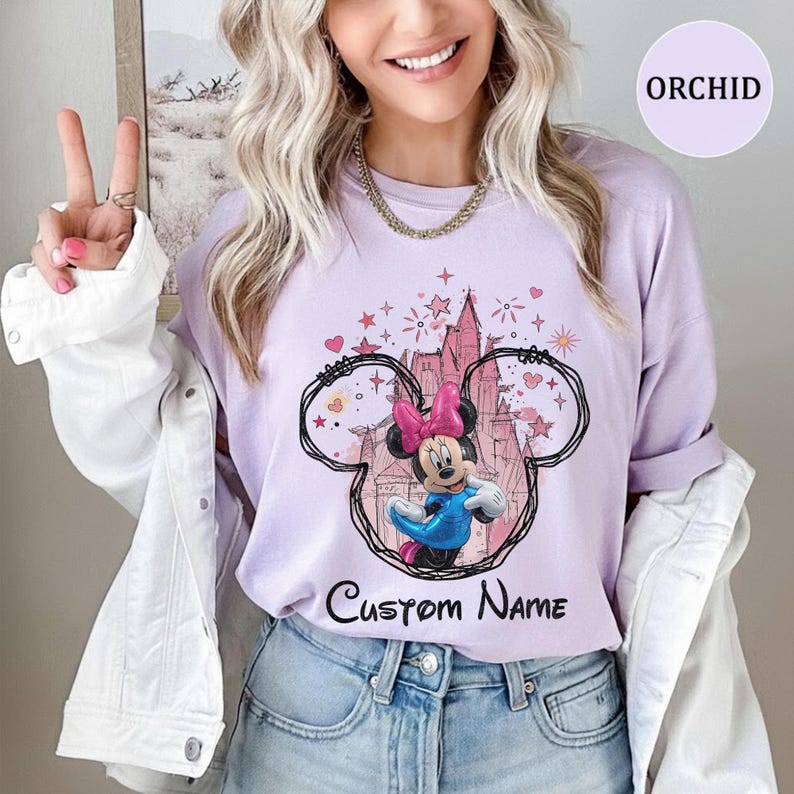 May include: A light purple t-shirt featuring a graphic of Minnie Mouse wearing a blue dress and a pink bow. The design includes a black outline of Mickey Mouse ears, a pink castle silhouette, stars, hearts, and the text Custom Name in black.