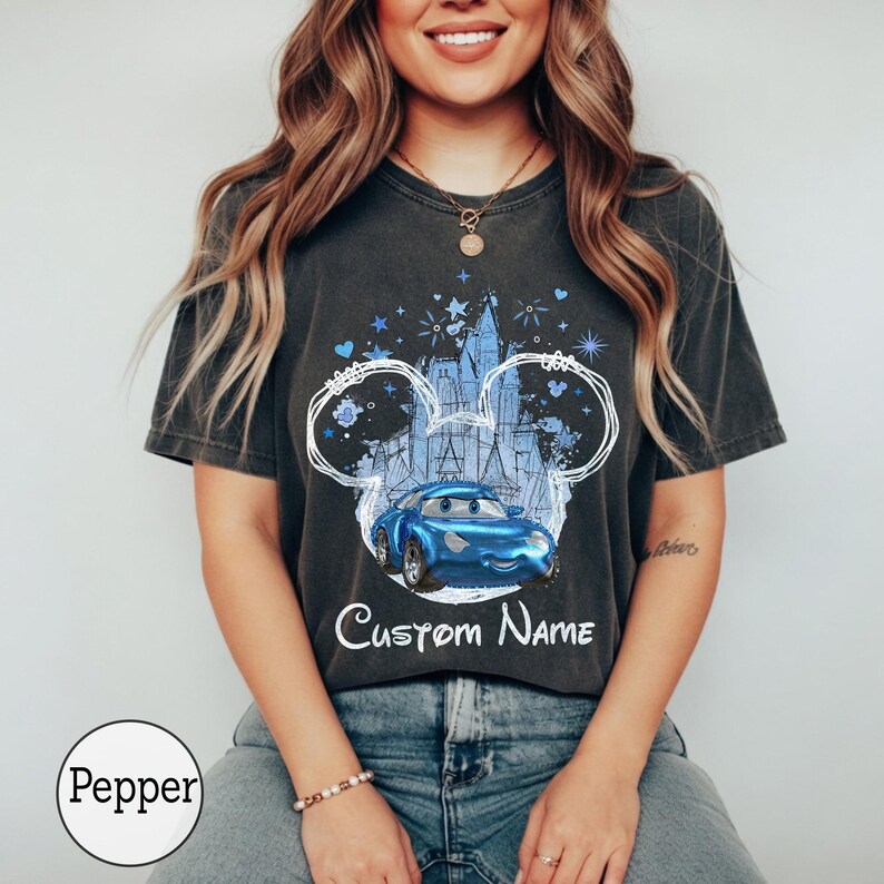 K&ouml;nnte beinhalten: Ein anthrazitfarbenes Kurzarm-T-Shirt mit einer Grafik eines blauen Cartoon-Rennwagens, zentriert in einer wei&szlig;en Umrissform von Mausohren. Hinter dem Auto befindet sich eine hellblaue Schlossillustration mit Sternen und Herzen. Der Text Custom Name steht darunter.