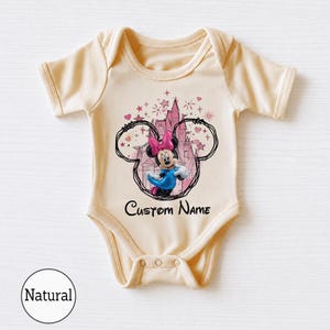 May include: A light beige short-sleeve baby bodysuit featuring a graphic of Minnie Mouse in a blue dress and pink bow, framed by a black outline of Mickey ears and a pink castle. The text Custom Name is printed in black below the graphic.