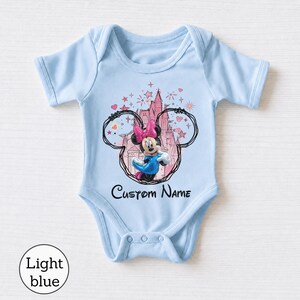 May include: A light blue baby bodysuit featuring a graphic of Minnie Mouse inside a black outline of Mickey Mouse ears. Behind the character is a pink castle illustration with stars and hearts. The text Custom Name is printed in black below.