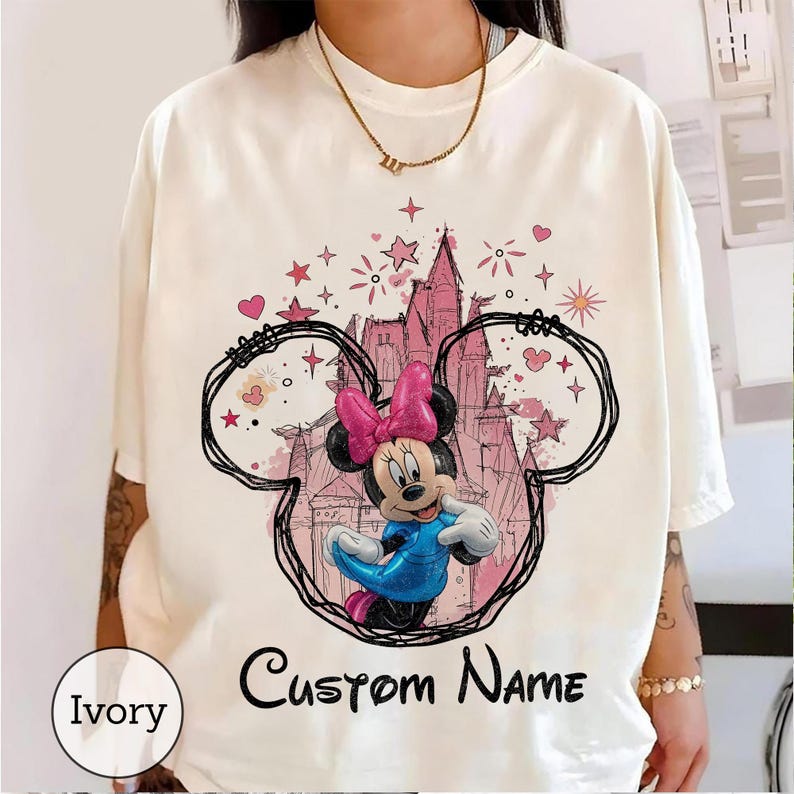 May include: An ivory short-sleeve t-shirt featuring a graphic of Minnie Mouse in a blue dress and pink bow, framed by a black hand-drawn ear outline. Behind the character is a pink castle illustration with stars and hearts. The text Custom Name is printed below.