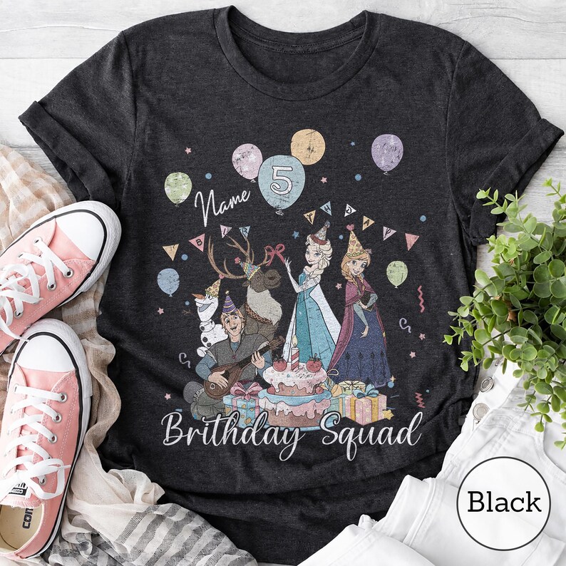 Op de afbeelding: Een donkergrijs t-shirt met een grafische print van geanimeerde figuren, waaronder een sneeuwpop, een rendier en twee figuren in jurken, omringd door ballonnen, een verjaardagstaart en cadeaus. De tekst luidt Name, 5 en Birthday Squad.
