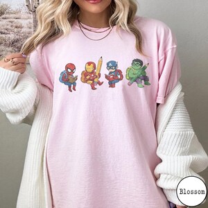 May include: A light pink t-shirt featuring a graphic print of four cartoon superheroes: Spider-Man reading a book, Iron Man holding a large yellow pencil, Captain America reading a book, and the Hulk holding a green pencil, all wearing backpacks.