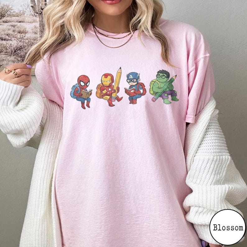 May include: A light pink t-shirt featuring a graphic print of four cartoon superheroes: Spider-Man reading a book, Iron Man holding a large yellow pencil, Captain America reading a book, and the Hulk holding a green pencil, all wearing backpacks.