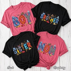 May include: Four short-sleeve t-shirts in black and raspberry pink featuring graphic prints of Disney Pixar Cars characters. The designs include the words BOY, BROTHER, SISTER, and MOM, decorated with checkered patterns and race car motifs.