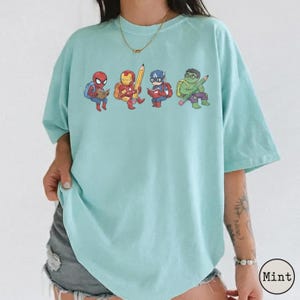 May include: A mint green short-sleeve t-shirt featuring a graphic print of four cartoon superheroes, including Spider-Man, Iron Man, Captain America, and Hulk, each engaged in reading or writing with school supplies like books and a large pencil.