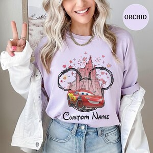 K&ouml;nnte beinhalten: Ein hellviolettes T-Shirt mit Rundhalsausschnitt und einer Grafik eines roten Rennwagens vor einer Schloss-Silhouette, umrahmt von einer schwarzen Umrisslinie von Mausohren, mit Sternen, Herzen und dem Text Custom Name in schwarzer Schrift.