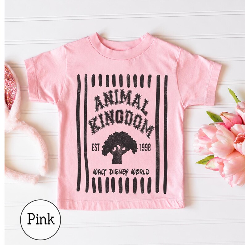 Pu&ograve; includere: Una t-shirt a maniche corte rosa chiaro con una stampa grafica nera. Il design include strisce verticali, il testo "ANIMAL KINGDOM", "EST 1998" e "WALT DISNEY WORLD", insieme alla silhouette centrale di un grande albero.