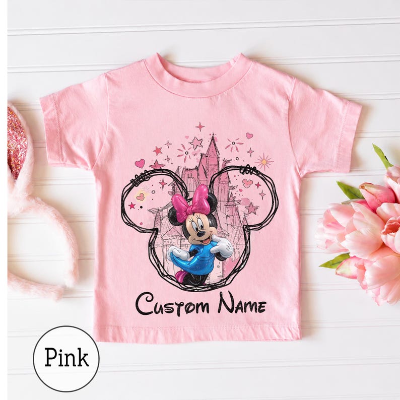 May include: A light pink short-sleeved t-shirt featuring a graphic of Minnie Mouse in a blue dress and pink bow, framed by a black hand-drawn mouse ear outline. Behind the character is a castle illustration with stars and hearts, and the text Custom Name.